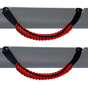 Puede incluir: Dos agarraderas para vehículo con un diseño de paracord trenzado en colores negro y rojo alternados. Cada agarradera está sujeta a una barra de montaje gris con herrajes negros en ambos extremos, creando un agarre curvo y texturizado.