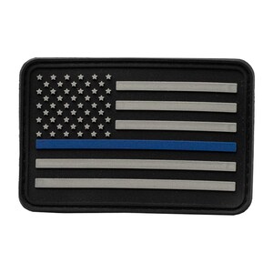 May include: A rectangular black patch featuring a stylized American flag design. The flag includes white stars on a black field, gray stripes, and a single blue stripe. The patch has a raised, textured border.