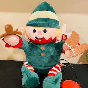 Puede incluir: Un muñeco de elfo navideño de peluche con gorro y atuendo verdes, ribetes rojos y piernas a rayas. El elfo sostiene galletas de jengibre y tiene una cara sonriente. Un pedal rojo está conectado al muñeco.