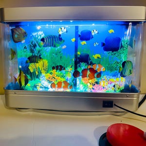 May include: A rectangular aquarium with a silver frame and a blue interior. The tank is filled with colorful artificial coral and various fish, including clownfish and angelfish. A black power switch is visible on the front.