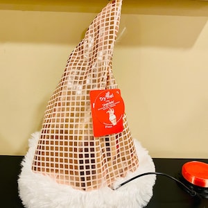 May include: A festive Santa hat with a shimmering, square-patterned gold fabric. The hat has a fluffy white trim and a red tag that says "Try me!". A black wire and red button are visible.
