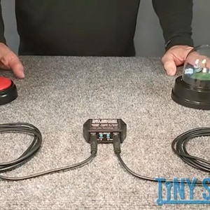 Puede incluir: Un botón rojo y un pequeño globo de nieve están conectados a una caja de control negra con cables negros. El globo de nieve se encuentra sobre una base negra. El texto "Tiny Switchy" está en la parte inferior derecha.