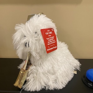 Puede incluir: Un juguete de cachorro animado blanco y esponjoso con una etiqueta roja que dice "¡Pruébame!". La etiqueta enumera tres canciones navideñas. El juguete tiene un collar dorado y está sobre una superficie negra.