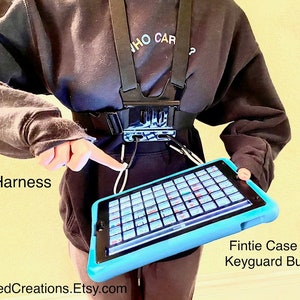 以下が含まれることがあります： 「WHO CAR?」と書いてある黒いスウェットシャツを着た人が、黒いチェストハーネスを着用し、タブレットを装着しています。タブレットは青いケースに入っていて、画面にはカラフルな正方形のグリッドが表示されています。画像の左側には「AAC Harness」、右側には「Fintie Case and Keyguard Bundle」というテキストが表示されています。画像の下部には「AdaptedCreations.Etsy.com」というテキストが表示されています。