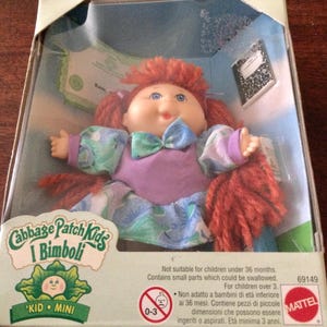 May include: A Cabbage Patch Kids "Kid Mini" doll in its original packaging. The doll has red yarn hair, blue eyes, and is wearing a purple dress with a blue bow. The box features the Cabbage Patch Kids logo and a warning label.