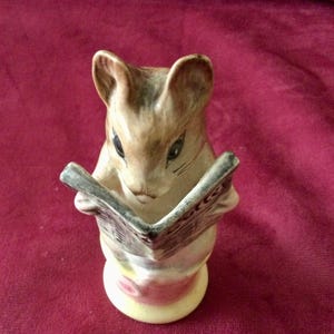 May include: A ceramic figurine of a brown and cream-coloured mouse holding an open newspaper. The mouse has pointed ears, dark eyes, and whiskers, with the newspaper featuring printed text and lines. The base is a rounded, light-coloured pedestal.