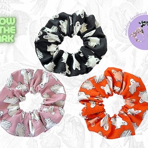 Op de afbeelding: Drie Halloween-scrunchies in zwart, roze en oranje, elk met een patroon van witte spookfiguren. De scrunchies worden weergegeven tegen een lichte achtergrond met de tekst "Glow in the Dark" en een afbeelding van bijpassende haarbanden.