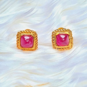 May include: A pair of gold earrings with a square shape and a red gemstone in the center. The earrings have a rope-like design around the edge.