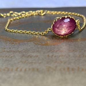 May include: A gold chain bracelet featuring a pink star ruby gemstone. The ruby is set in a gold bezel with a scalloped edge.