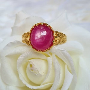 May include: A gold ring with a large, oval-shaped, pink ruby gemstone set in a decorative, gold setting. The ring is on a white background.