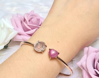 May include: A gold bangle bracelet with a rose quartz and a pink heart shaped gemstone. The bracelet is on a person's wrist.
