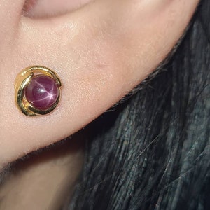 Op de afbeelding: Een paar gouden oorbellen met een rode stervormige edelsteen in het midden. De oorbellen zijn simpel van ontwerp met een gebogen vorm.