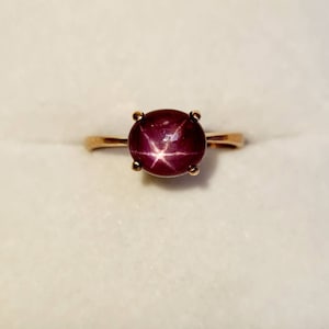 Peut inclure: Une bague en or rose ornée d'une grosse pierre précieuse ronde, rouge foncé, avec un motif en étoile à six branches. La pierre est maintenue par quatre griffes. La bague est posée sur un fond blanc.