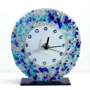 Può includere: Un orologio decorativo in vetro con quadrante bianco e lancette nere. Il quadrante è circondato da pezzi di vetro blu e turchese. L'orologio poggia su una base rettangolare blu scuro.