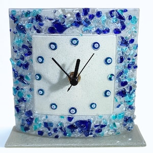 Può includere: Un orologio quadrato in vetro con un quadrante bianco con disegni blu a forma di occhio che segnano le ore. La cornice dell'orologio è decorata con pezzi di vetro blu e turchese. L'orologio ha lancette nere e un centro color bronzo.