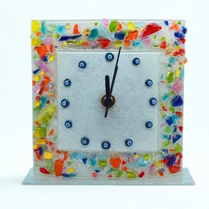 Può includere: Un orologio quadrato in vetro con quadrante bianco e lancette nere. Il quadrante ha indicatori blu a forma di occhio. La cornice è decorata con pezzi di vetro colorati. L'orologio misura circa 10 cm di larghezza e 13 cm di altezza.