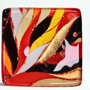 以下が含まれることがあります： 鮮やかな抽象的なデザインの正方形のガラスアート作品。赤、黒、黄、白、金などの色が混ざっています。デザインは、流れるような有機的な形とパターンで構成されています。