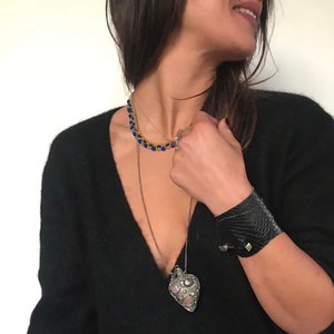Könnte beinhalten: Eine Frau trägt einen schwarzen Pullover mit einer silbernen herzförmigen Halskette mit rosa und weißen Steinen. Sie trägt auch eine blaue Perlenkette und ein schwarzes Lederarmband.