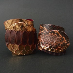 Könnte beinhalten: Zwei Lederarmbänder mit Schlangenhautmuster. Ein Armband ist gold und braun, das andere ist braun und rot.