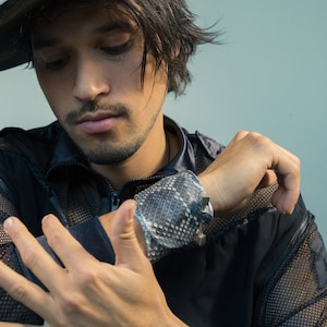 Peut inclure: Un homme portant une chemise en maille noire avec un bracelet en peau de serpent et un chapeau à large bord avec une finition blanche vieillie.