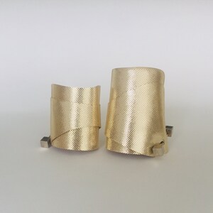 May include: Two gold metal bracelets with a textured surface. The bracelets are different sizes and are stacked on top of each other.