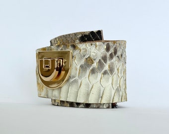 Könnte beinhalten: Ein weißes und braunes Schlangenlederarmband mit einem goldenen Verschluss. Das Armband ist aus Leder und hat eine strukturierte Oberfläche.