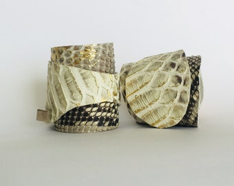 Könnte beinhalten: Zwei weiße Lederarmbänder mit goldenen und schwarzen Akzenten. Die Armbänder sind aus Schlangenleder und haben eine strukturierte Oberfläche.