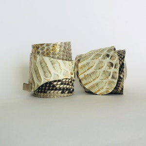 Könnte beinhalten: Zwei weiße Lederarmbänder mit goldenen und schwarzen Akzenten. Die Armbänder sind aus Schlangenleder und haben eine strukturierte Oberfläche.