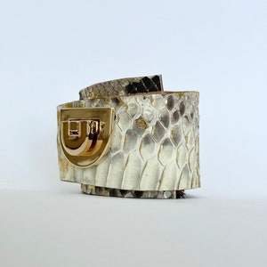 Könnte beinhalten: Ein weißes und braunes Schlangenlederarmband mit einem goldenen Verschluss. Das Armband ist aus Leder und hat eine strukturierte Oberfläche.