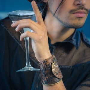 Könnte beinhalten: Eine Person trägt ein schwarzes Netzhemd mit einem braunen Lederarmband. Das Armband hat ein Schlangenhautmuster. Die Person hält ein Martini-Glas.
