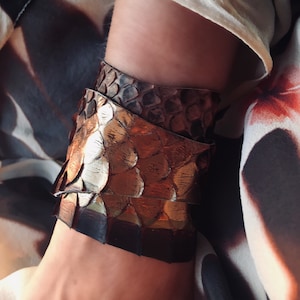 May include: A brown leather bracelet with a snakeskin pattern. The bracelet is wrapped around a person's wrist.