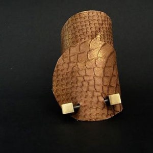 May include: A brown leather cuff bracelet with a textured snakeskin pattern and two gold square accents.