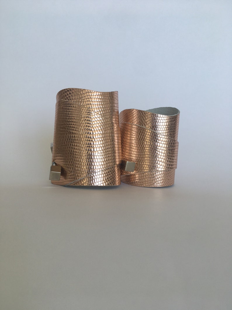 May include: Two rose gold leather bracelets with a textured finish. The bracelets have a silver metal clasp.