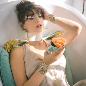 Könnte beinhalten: Eine Frau in einem weißen Hemd mit Kragen und einer beigefarbenen Hose sitzt in einer Badewanne und hält eine hellblaue Teetasse mit einer pfirsichfarbenen Rose darin. Sie trägt ein gold-schwarz gemustertes Manschettenarmband am rechten Handgelenk und ein braun-schwarz gemustertes Manschettenarmband am linken Handgelenk.