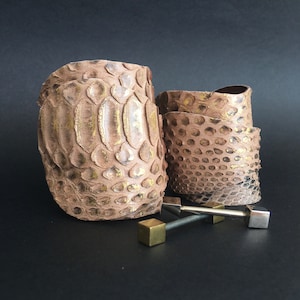 Könnte beinhalten: Zwei braune Lederarmbänder mit Schlangenhautmuster und goldenen Akzenten. Die Armbänder sind übereinander gestapelt und werden von zwei Metallverschlüssen begleitet.