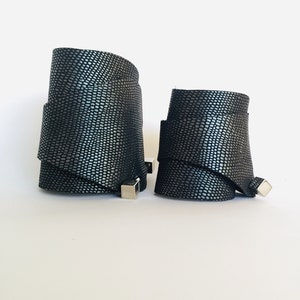 Op de afbeelding: Twee zwarte leren armbanden met een zilverkleurige gesp. De armbanden hebben een getextureerd, schubbig patroon.