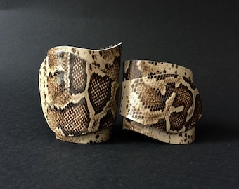 Könnte beinhalten: Zwei braune und weiße Lederarmbänder mit Schlangenhautmuster. Die Armbänder sind beide breit und haben eine leicht gekrümmte Form.