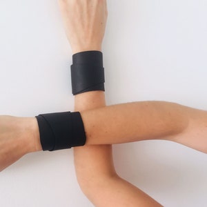 May include: Two black leather bracelets on a person's wrists. The bracelets are wide and wrap around the wrists.