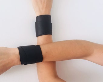 May include: Two black leather bracelets on a person's wrists. The bracelets are wide and wrap around the wrists.