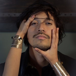 May include: A person with dark hair is wearing a black leather jacket and has their hand on their chin. They are wearing a gold bracelet on their wrist.