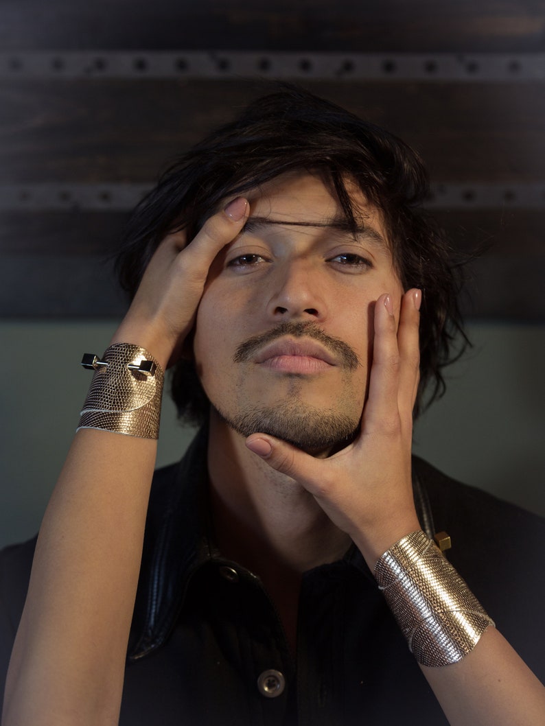 May include: A person with dark hair is wearing a black leather jacket and has their hand on their chin. They are wearing a gold bracelet on their wrist.