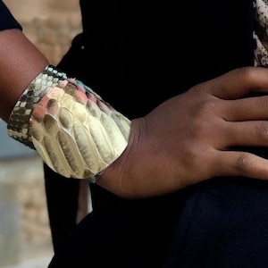 Könnte beinhalten: Ein breites, goldfarbenes Armband mit einem strukturierten, schlangenhautartigen Muster. Das Armband ist aus Leder und hat ein mehrfarbiges Design.