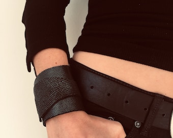 Puede incluir: Un brazalete de cuero negro con una superficie texturizada. El brazalete se lleva en la muñeca de una persona que lleva una camisa negra y unos vaqueros negros.