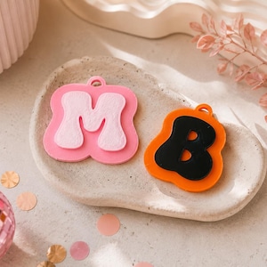 May include: Two layered acrylic letter charms, one pink with a white "M" and the other orange with a black "B". The charms are on a light-colored, textured tray, with a disco ball and confetti in the background.