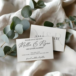 May include: Two white wedding announcement cards with black text, one reading "Mollie & Ryan ARE GETTING MARRIED!" and the other "SAVE THE DATE". Eucalyptus leaves are arranged around the cards on a white fabric background.