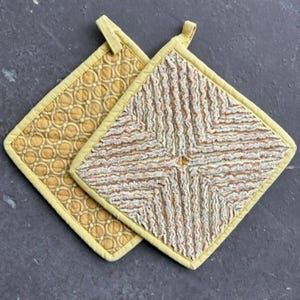 May include: Two square pot holders with yellow trim. One has a repeating pattern of yellow circles, while the other features a textured, striped design in cream and brown. Both have a loop for hanging.
