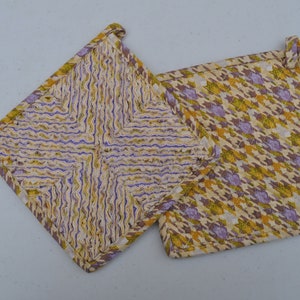 May include: Two square pot holders with a quilted design. One features a pattern of yellow and purple wavy lines, while the other has a houndstooth pattern in yellow, purple, and white. Both have a loop for hanging.