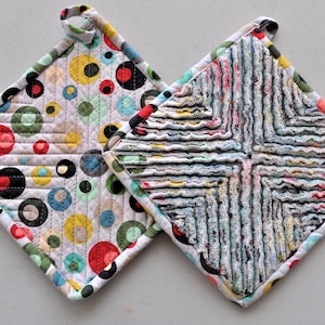 May include: Two colorful pot holders, one with a geometric pattern of circles and the other with a woven pattern of strips of fabric.