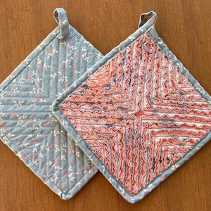 May include: Two quilted pot holders. One is blue with a floral pattern and the other is pink with a ruffled design.