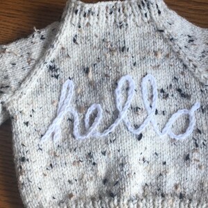Könnte beinhalten: Cremefarbener Strickpullover mit schwarzen, braunen und beigen Flecken. Das Wort "hello" ist mit weißem Garn auf die Vorderseite gestickt. Der Pullover hat einen gerippten Halsausschnitt und Bündchen und ist wahrscheinlich für ein Baby oder Kleinkind.